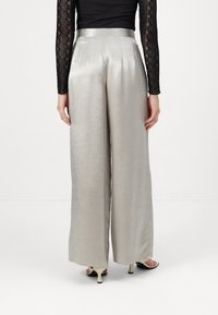 Wide-leg trousers in shiny silver fabric, featuring a high waist, sharp pleats, and a smooth texture. Styled with black lace sleeves.