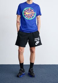 Modré tričko s kruhovým grafickým motivem a neonovými akcenty, doplněné o černé kraťasy s bílým nápisem a logem Nike; černé ponožky a tenisky.