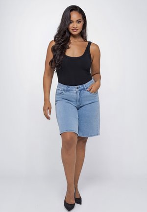 Woman with long dark hair wearing a black tank top, light blue denim shorts, and black heels, standing against a plain background.