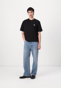 Svart bomulls T-skjorte med korte ermer og en liten logo-patch. Brukt sammen med lyseblå løs-passform jeans og svarte sko, stående foran en hvit bakgrunn.