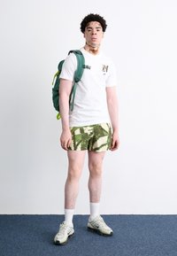 Witte T-shirt met een groene rugzak, groen-witte patronen shorts, witte sokken en lichtgrijze sneakers met zwarte accenten, staand op een tapijt.