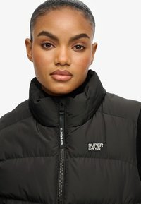 Chaleco acolchado negro con cuello alto, cierre de cremallera y parche con el logo en el pecho izquierdo. Textura lisa y brillante, típica de prendas ligeras de exterior para invierno.