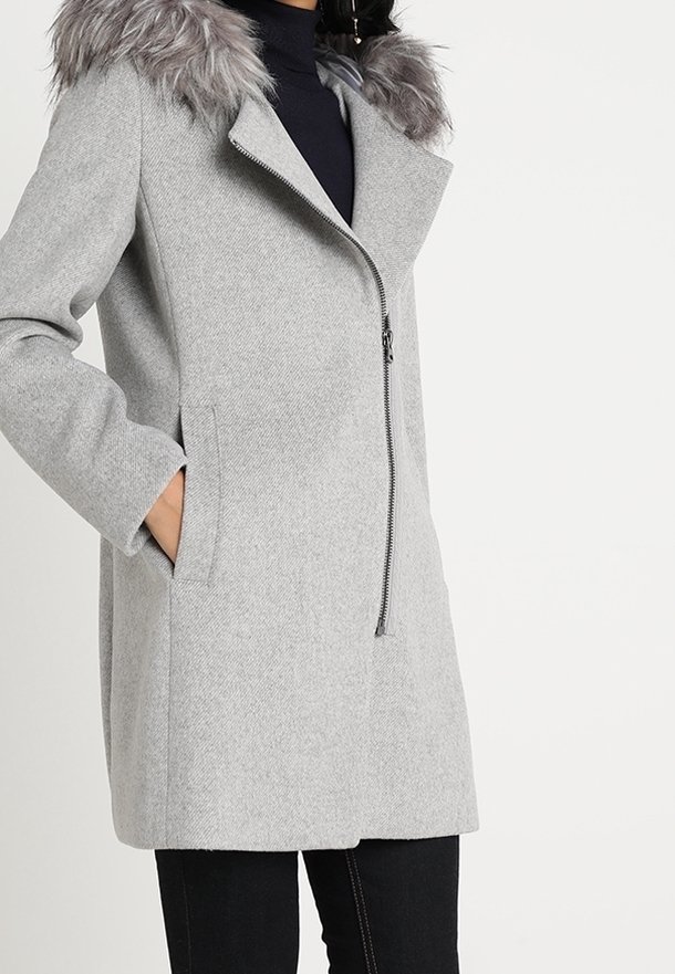 Femme portant un manteau en laine gris clair avec une fermeture éclair asymétrique et un col en fausse fourrure grise, mains dans les poches latérales.