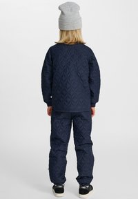 Dziecko ubrane w granatową pikowaną kurtkę i spodnie, szarą czapkę oraz czarne trampki, stojące tyłem na jednolitym białym tle.