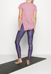 Woman standing barefoot on black yoga mat wearing pink short-sleeve top and blue leopard-print leggings, shown from behind.