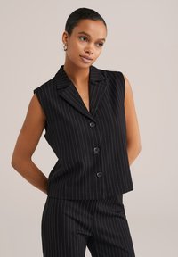 Černá bezručková vesta s vertikálními bílými pruhy, strukturovaným límcem a pěti černými knoflíky. K tomu ladící kalhoty s proužky dokončují outfit.
