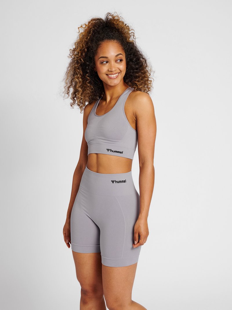 Jonge vrouw met krullend haar, draagt een bijpassende lichtgrijze Hummel sportbeha en hoogtallige biker shorts, staand en glimlachend, kijkend naar links.