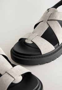 Sandales en daim gris clair avec semelle en caoutchouc noire, présentant des brides croisées et un design à bout arrondi. Surface texturée et semelle épaisse.