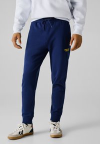 Navy Jogginghosen aus weichem Stoff, mit elastischem Bund und Kordelzug, seitlichen Taschen und einem gelben Logo am linken Oberschenkel.