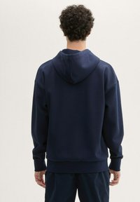 Granatowy hoodie o miękkiej fakturze, z kapturem na sznurki, ściągaczami przy mankietach i dole oraz luźnym krojem. Brak widocznych wzorów lub akcentów.