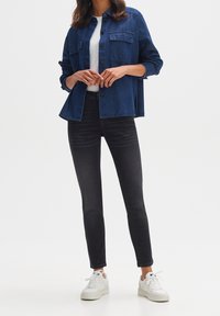 Femme portant une veste en jean bleu foncé par-dessus un haut blanc, un jean skinny noir et des baskets blanches, debout devant un fond uni.