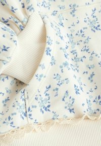 Le tissu à motif floral présente de petites fleurs bleues sur un fond crème, avec des accents côtelés doux et une délicate bordure en dentelle.