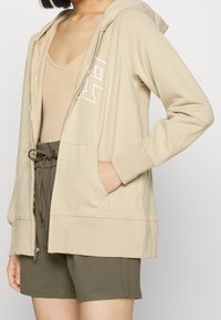 Beige mikina na zip s kangaroo kapsami a bílým logem, v kombinaci s olivově zelenými šortkami pro uvolněný vzhled. Měkký materiál a volný střih.