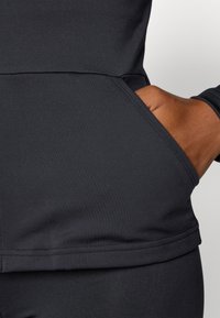 Black athletic top featuring a side pocket and smooth, stretchy fabric. The design includes a fitted shape and a contrasting seam detail.
