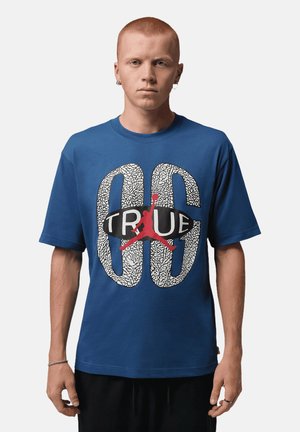Jonge man met kort rood haar die een blauw T-shirt draagt met een grote witte en zwarte gebarsten print en een rood basketbalspelerlogo.
