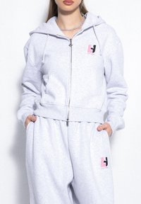 Szara bluza z kapturem na zamek błyskawiczny i sznurkiem, z małym logo w kolorze różowym i czarnym. Wykonana z miękkiej, teksturowanej tkaniny. Uzupełnia dopasowane dresy.