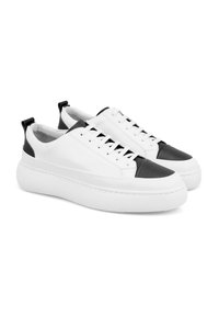 Witte leren sneakers met een zwarte rubberen neuskap, dikke witte zool en zwarte accenten op de hiel. Vetersluiting, gladde textuur.