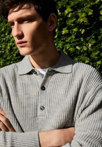 Hellgraues Baumwollstrick-Polohemd mit einem gerippten Muster, drei schwarzen Knöpfen und einem Kragen. Das Model steht mit verschränkten Armen vor einem grünen Hintergrund.