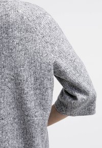 Manteau en laine grise texturée à manches trois quarts porté par une personne sur un fond blanc uni.