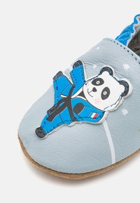 Zapatilla de cuero azul claro con un gráfico de un panda vestido de azul. Suela suave con sutiles detalles texturizados y estrellas impresas alrededor del panda.