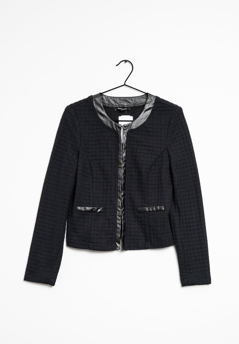 Veste noire texturée avec un col rond, bordure noire brillante, fermeture éclair et deux poches avant. Suspendue à un crochet sur un fond blanc.