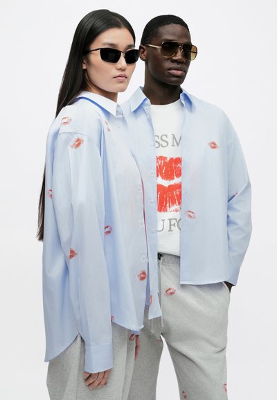 Light blue button-up shirt with red kiss print accents, worn over a graphic t-shirt and gray sweatpants. Black and gold sunglasses.