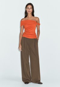 Oranger, schulterfreier Oberteil mit figurbetonter Passform; kombiniert mit braunen, weit geschnittenen Hosen und schwarzen Schuhen. Der Hintergrund ist schlicht und hell.