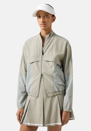 Kobieta w beżowej kurtce na zamek z wzorzystymi rękawami i panelami, dopasowanej plisowanej spódnicy i białej daszku, stojąca na jednolitym tle.