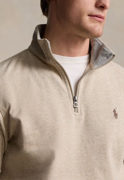 Hombre con sudadera beige con cremallera hasta un cuarto, encima de camiseta blanca, mostrando detalle de la cremallera y pequeño logo bordado en el pecho.