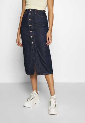 Mujer con falda midi vaquera azul oscuro con botones delanteros, zapatillas blancas gruesas con cordones y top de rayas amarillo claro.