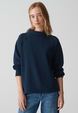 Felpa blu navy con collo alto, maniche raglan e polsini a coste. Texture liscia e vestibilità rilassata, abbinata a jeans azzurri.