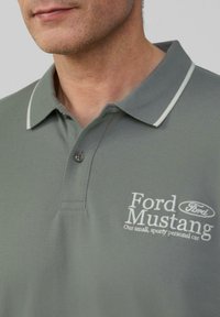 Man draagt een grijze polo met witte accenten en een wit "Ford Mustang" logo en tekst op de borst.