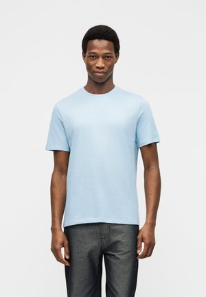 Egy fiatal férfi áll szemben velünk világoskék rövid ujjú pólót és sötétszürke nadrágot viselve, egyszínű fehér háttér előtt.