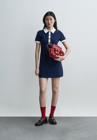 Young woman in navy blue dress with white collar and cuffs holds red handbag, wearing red socks and black loafers, standing on gray floor.