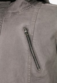 Grau Jacke mit strukturiertem Stoff, einer markanten silbernen Reißverschluss-Tasche und einer Kapuze, sowie Nähte an den Schultern.