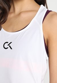 Top deportivo atlético blanco con un logo negro "CK", superpuesto sobre un sujetador deportivo color burdeos. Textura suave con tirantes delgados y escote redondo.
