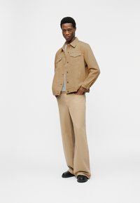 Άνδρας που στέκεται με τα χέρια στις τσέπες, φοράει μπεζ μπουφάν και μπεζ παντελόνι φαρδιάς γραμμής με μαύρα παπούτσια, σε λευκό φόντο.