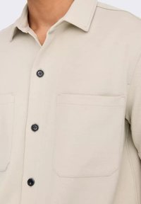 Camisa de botón beige claro que presenta un cuello, dos bolsillos en el pecho y botones negros, fabricada con un tejido texturizado.