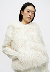 Mulher vestindo um suéter grosso branco, sobreposto a um colete fofinho e texturizado em off-white, de pé contra um fundo claro e simples.