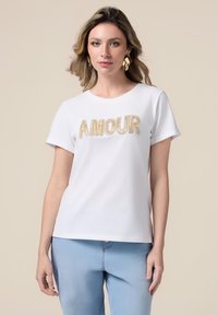 Camiseta de algodón blanca con la palabra "AMOUR" en hilo dorado, de mangas cortas y cuello redondo, combinada con jeans de mezclilla azul claro.