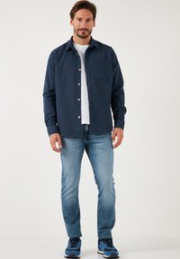 Camicia button-up navy con taschino sul petto, indossata sopra una maglietta bianca, abbinata a jeans azzurri e sneakers blu. Tessuto della trama liscia.