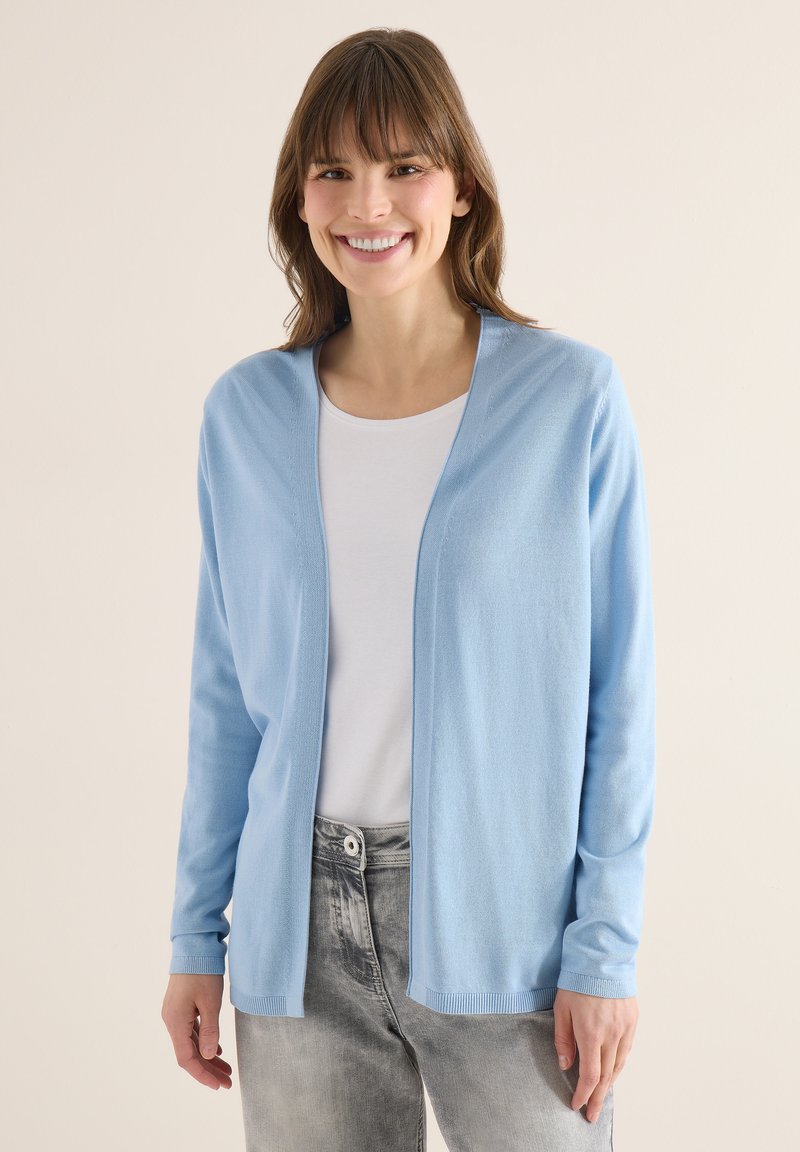 Femme souriante portant un cardigan bleu clair ouvert, une chemise blanche et un jean gris, sur un fond beige uni.