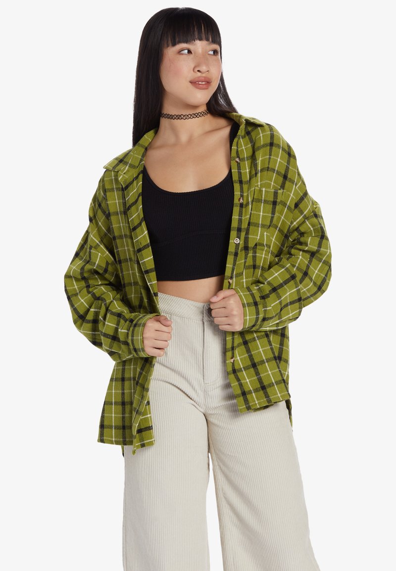 Zielona flanelowa koszula w kratę z zapięciem na guziki, w oversize'owym fasonie i czarnymi wykończeniami. Noszona na czarnym topie typu crop i kremowych spodniacjach o szerokich nogawkach.