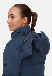 Marineblauwe gewatteerde jas met een hoge kraag, zichtbare stiksels en een zwart logo-opdruk op de mouw. Beschikt over een open capuchon met trekkoord.