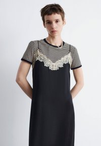 Schwarzes Kleid mit Spitzenverzierungen über einem gestreiften Hemd. Das Kleid hat eine fließende Silhouette und eine weiche Textur, mit kurzen Ärmeln und einem runden Ausschnitt.