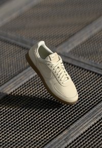 Bežiniai Nike sportbačiai su lygaus odos viršumi, apvaliu pirštu ir guminiu padu, pakabinti virš tekstūruoto paviršiaus.