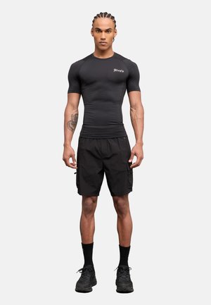 Hombre atlético con cabello trenzado de pie mirando hacia adelante, vistiendo una camiseta negra ajustada de manga corta, pantalones cortos cargo negros, calcetines negros y zapatillas negras.