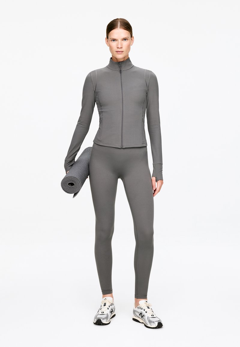 Tenue de sport grise comprenant une veste zippée ajustée et un legging assorti, tous deux en tissu lisse et extensible. Le mannequin tient un tapis de yoga.