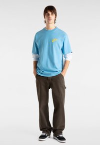 Vaaleansininen puuvillapaita, jossa on keltainen logo, kerrostettuna valkoisen pitkähihasen paidan päälle, yhdistettynä tummanruskeisiin housuihin ja mustiin tennareihin.