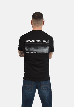 Uomo con capelli corti e tatuaggi indossa una maglietta nera Armani Exchange con scritta e grafica bianche, abbinata a jeans blu, di spalle.